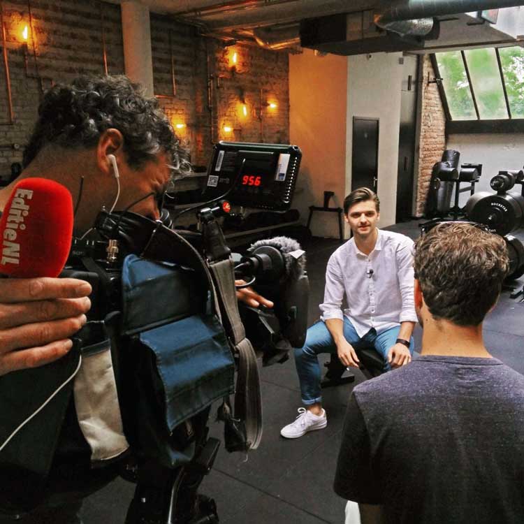 Man in white shirt and blue jeans sitting on a bench in a gym being interviewed on camera by a crew member holding a microphone and operator filming with professional video camera.
