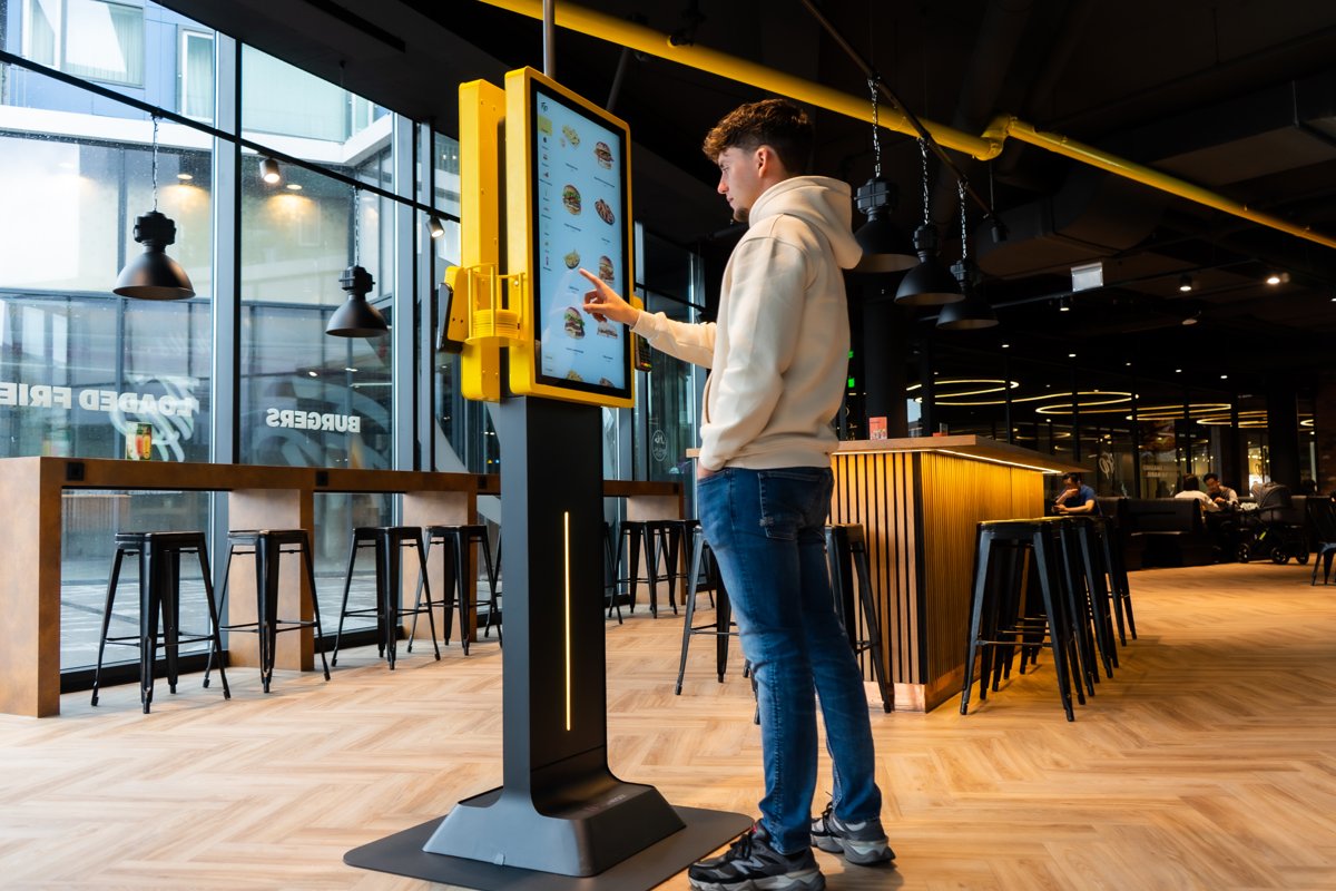 Guest ordering at a yellow Onesix Kiosk with a large touchscreen in a burger restaurant