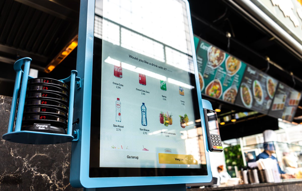 Onesix Kiosk Premium in a busy food hall, showing an English upsell prompt on the ordering screen and buzzers on the charging base