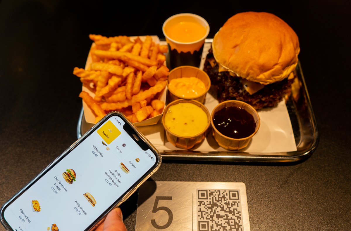 Guest browsing the digital menu on a phone next to a table plate with a table number and QR code, with a burger and fries on a tray