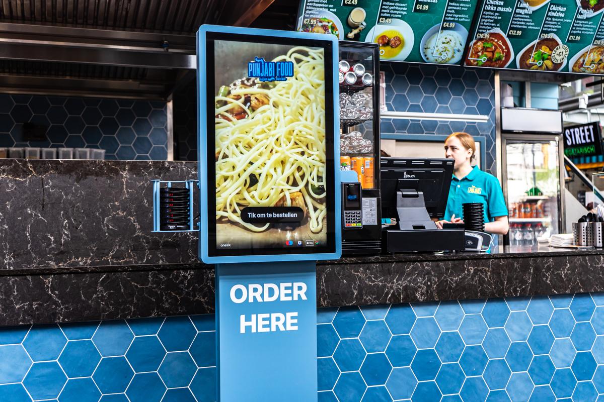 Blue Onesix Kiosk with 'Order here' next to the staffed counter in an Indian takeaway restaurant
